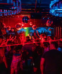 Crowded urban nightclub dance floor with mirrored disco balls, neon red and blue lighting and a green spotlight cutting through the crowd as people dance and cheer.