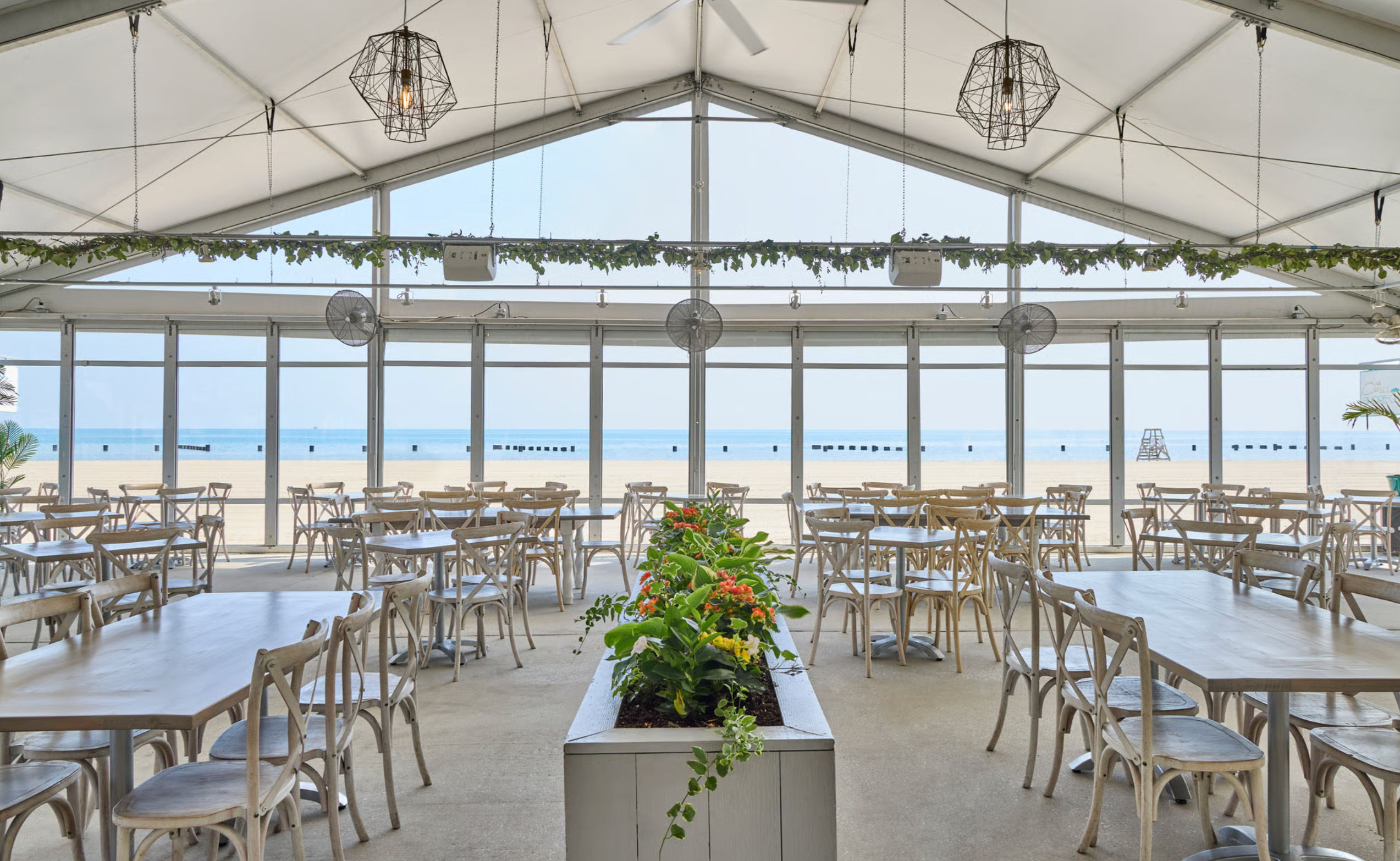 Sunlit beachfront dining tent with light wooden tables and cross-back chairs, a central planter of orange flowers, geometric pendant lights, and floor-to-ceiling windows framing a sandy beach and calm blue ocean.