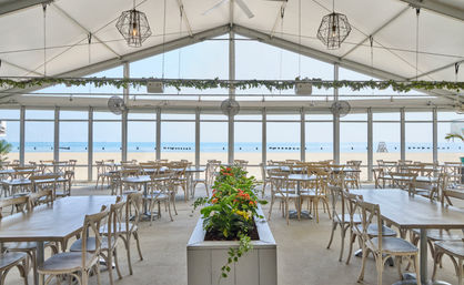 Sunlit beachfront dining tent with light wooden tables and cross-back chairs, a central planter of orange flowers, geometric pendant lights, and floor-to-ceiling windows framing a sandy beach and calm blue ocean.