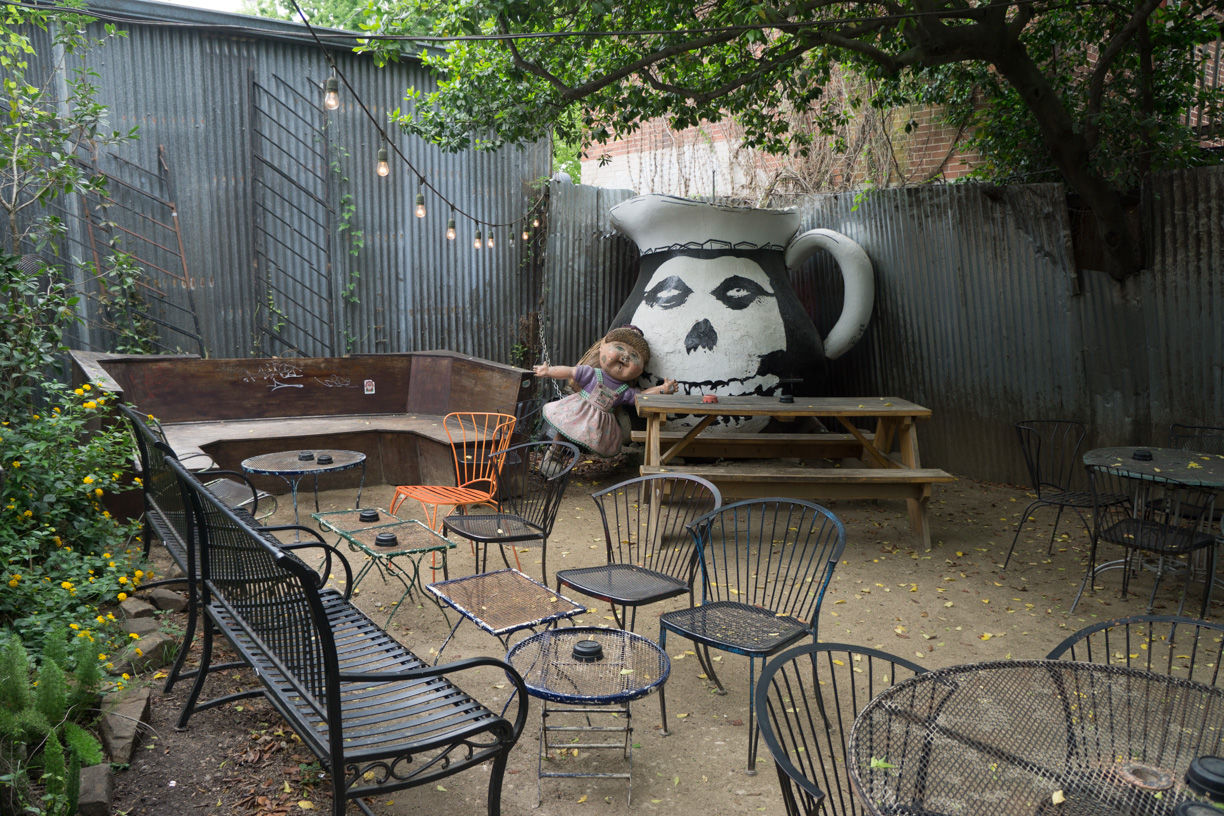 Outdoor garden patio with metal tables and chairs, picnic bench under string lights, corrugated metal fence, large painted skull-pitcher mural and an oversized doll on a swing.