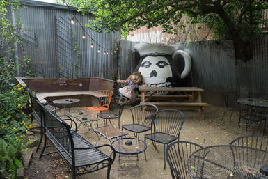 Outdoor garden patio with metal tables and chairs, picnic bench under string lights, corrugated metal fence, large painted skull-pitcher mural and an oversized doll on a swing.