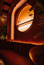 Cozy amber-lit lounge interior with a circular illuminated wall niche holding metal flamingo sculptures, plush curved banquette seating, tropical plants and warm wood accents.