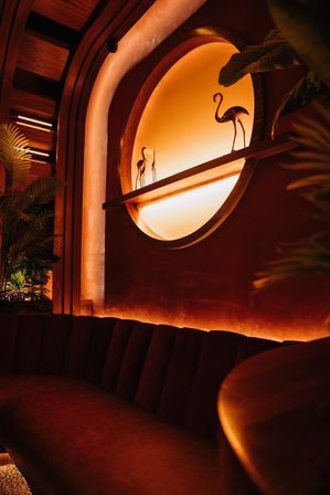 Cozy amber-lit lounge interior with a circular illuminated wall niche holding metal flamingo sculptures, plush curved banquette seating, tropical plants and warm wood accents.