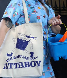 Person in a blue tropical shirt holding a canvas tote printed with a playful purple cartoon drink and the bold slogan 'ATTABOY', plus a blue sand pail and orange shovel — summer beach accessories
