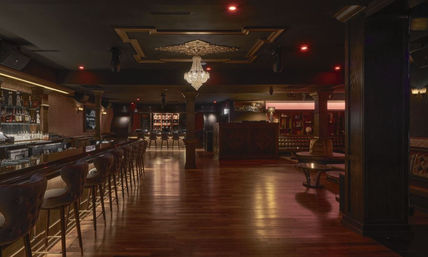 Moody upscale lounge bar interior with long wooden counter lined with leather bar stools, crystal chandelier, polished hardwood floor, and cozy booth seating under warm ambient lighting.