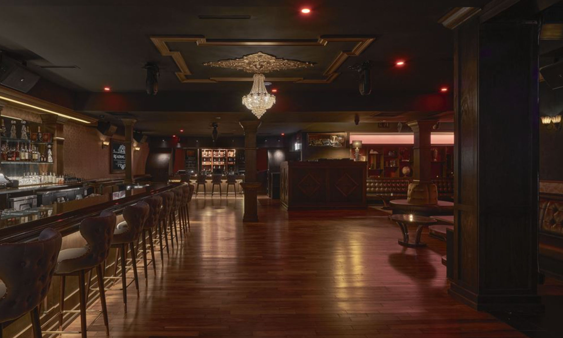 Moody upscale lounge bar interior with long wooden counter lined with leather bar stools, crystal chandelier, polished hardwood floor, and cozy booth seating under warm ambient lighting.