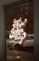 Retro script decal reading "Good Night John Boy" on a storefront window, warm brown tones and an urban street reflection with a parked car.