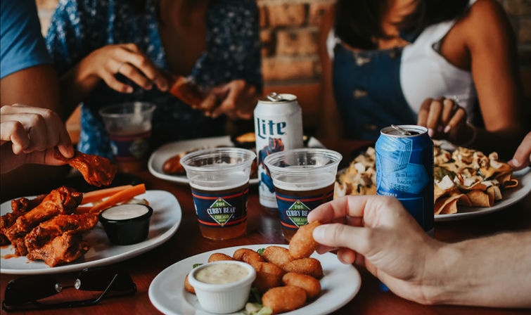Friends sharing bar snacks — saucy chicken wings, breaded cheese bites with dipping sauce, loaded nachos and beer cans on a wooden table.