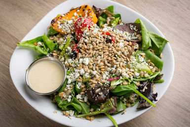 Fresh mixed greens salad in a white bowl with roasted red and yellow peppers, sautéed mushrooms, crumbled white cheese, a mound of sunflower seeds and a small metal cup of creamy dressing on a wooden table.