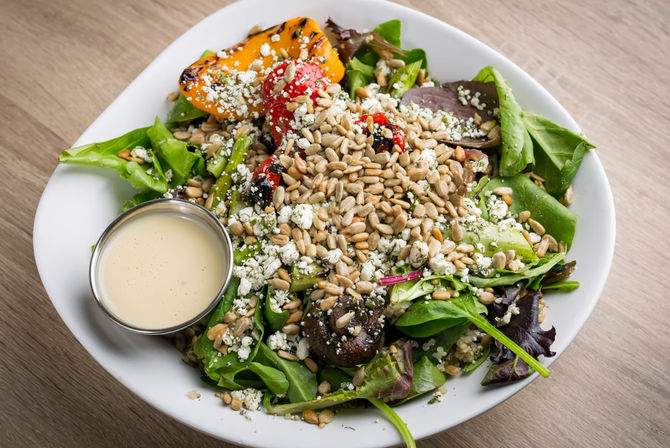 Fresh mixed greens salad in a white bowl with roasted red and yellow peppers, sautéed mushrooms, crumbled white cheese, a mound of sunflower seeds and a small metal cup of creamy dressing on a wooden table.