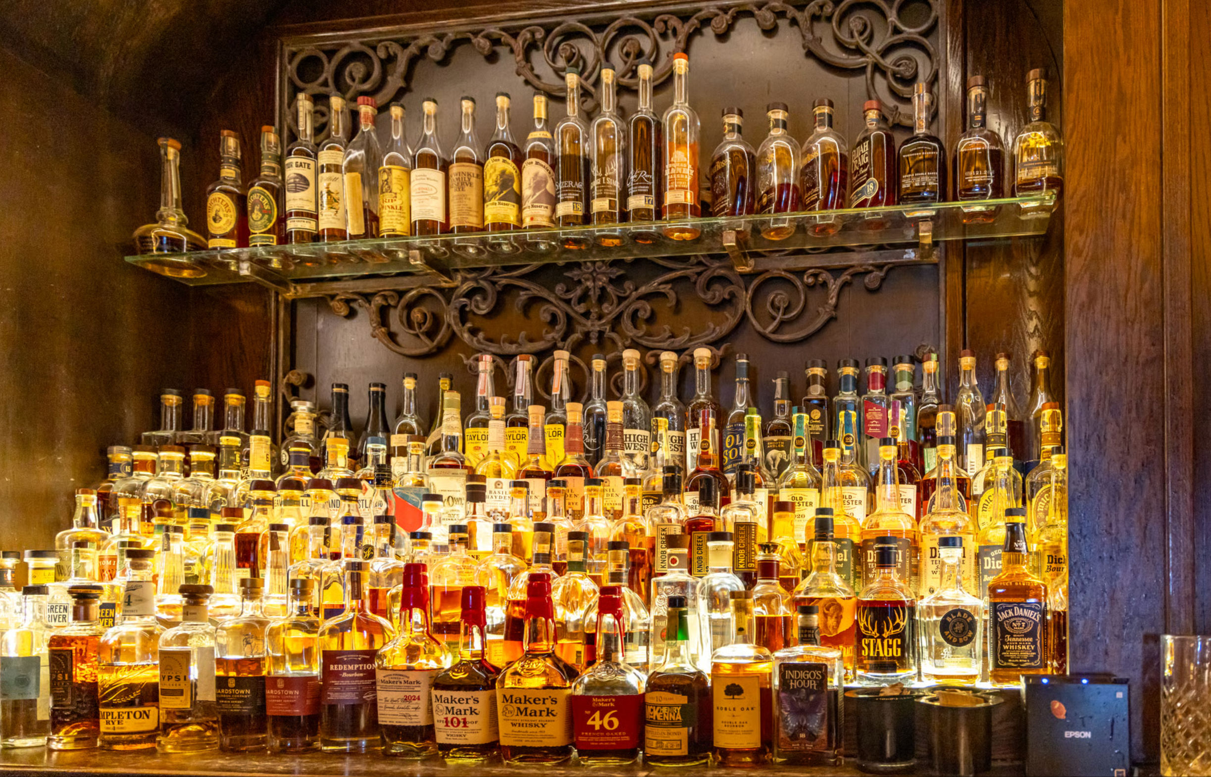 Glowing amber lineup of dozens of whiskey and bourbon bottles on glass shelves in a cozy back bar, framed by ornate dark wood and carved metal trim.