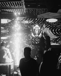Black-and-white nightclub scene with VIP bottle sparklers, lively crowd on the dance floor, and a large skull emblem on an LED wall.