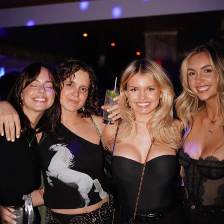 Four smiling women posing at a nightclub bar, dressed in black and holding cocktails under purple party lights — friends enjoying a night out.