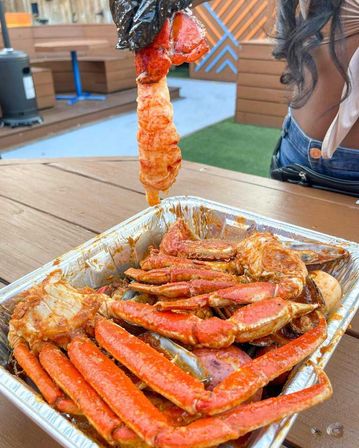 Aluminum tray of spicy seafood boil with bright red crab legs and a peeled shrimp held above a wooden outdoor patio table