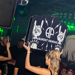 Woman holding a large black club sign with rock-style hand horns, lightning bolts and stylized initials, raised above a crowded nightclub dance floor under green disco lighting and a disco ball.
