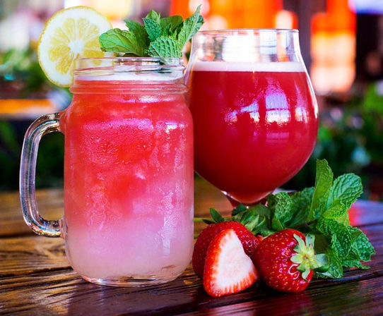 Two strawberry lemon drinks on a wooden table — a pink ombré mason-jar lemonade garnished with lemon slice and mint, alongside a deep-red strawberry beverage in a stemmed glass with fresh strawberries and mint, bright patio background.