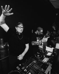 Black-and-white photo of two DJs in a crowded nightclub DJ booth — one with headphones raising his hand, the other adjusting the mixer and pointing, surrounded by turntables, CDJs and an energetic crowd.