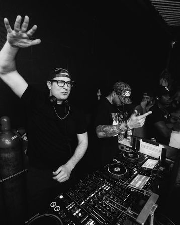Black-and-white photo of two DJs in a crowded nightclub DJ booth — one with headphones raising his hand, the other adjusting the mixer and pointing, surrounded by turntables, CDJs and an energetic crowd.