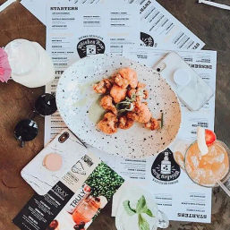 Overhead shot of a casual restaurant table with a speckled plate of crispy fried bites and garnish, menus underneath, a fruity cocktail with a strawberry, a mason-jar drink with mint, sunglasses and a phone
