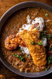 Bowl of New Orleans-style gumbo with white rice, grilled shrimp skewers, a crispy fried catfish fillet, and chopped green onions.