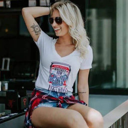 Smiling blonde woman in sunglasses sitting on a city cafe patio, wearing a white V-neck graphic tee, ripped denim shorts and a plaid shirt tied at her waist, showing arm tattoos — casual summer street style.