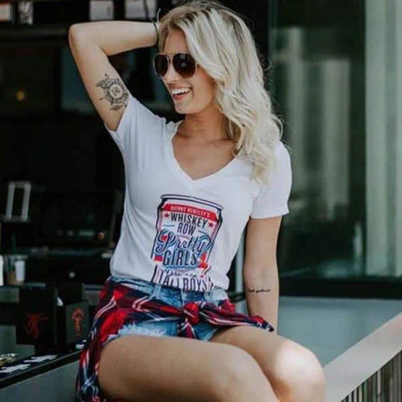 Smiling blonde woman in sunglasses sitting on a city cafe patio, wearing a white V-neck graphic tee, ripped denim shorts and a plaid shirt tied at her waist, showing arm tattoos — casual summer street style.