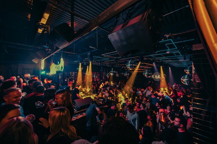 Packed nightclub dance floor at a late-night party, DJ booth and disco balls under warm stage lighting with a lively crowd dancing and socializing.