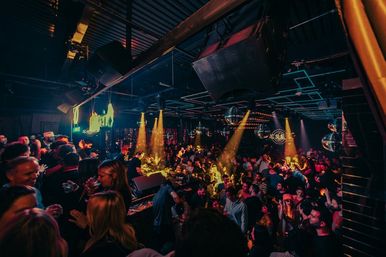 Packed nightclub dance floor at a late-night party, DJ booth and disco balls under warm stage lighting with a lively crowd dancing and socializing.