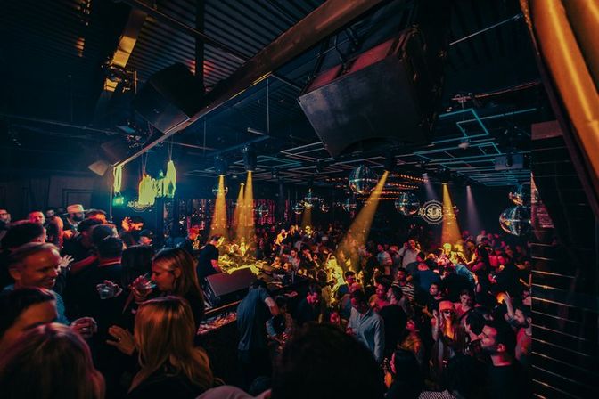 Packed nightclub dance floor at a late-night party, DJ booth and disco balls under warm stage lighting with a lively crowd dancing and socializing.