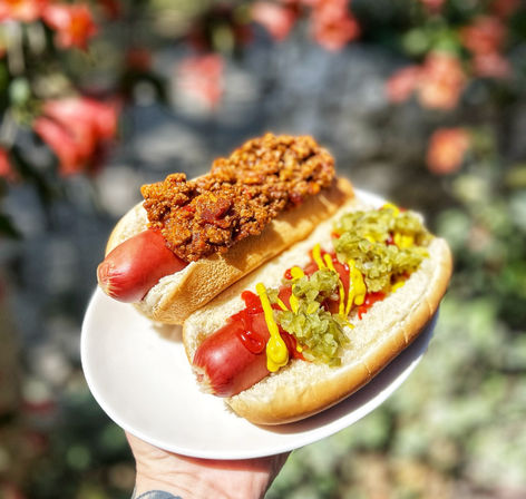 Close-up of two loaded hot dogs—one with chili, the other with mustard, ketchup and pickle relish—served on a white plate held in a sunny backyard garden.