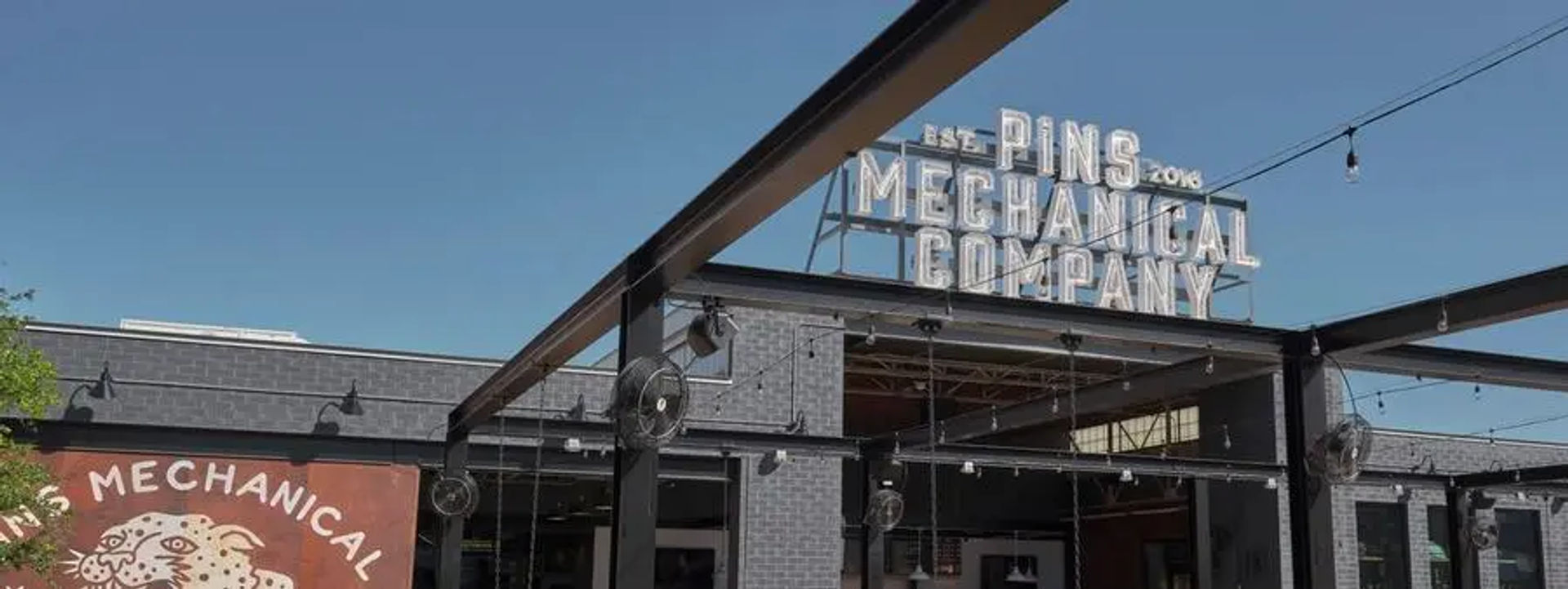 Industrial-chic outdoor patio with metal pergola, hanging string lights and fans, retro rooftop marquee sign atop a gray brick building under a clear blue sky.