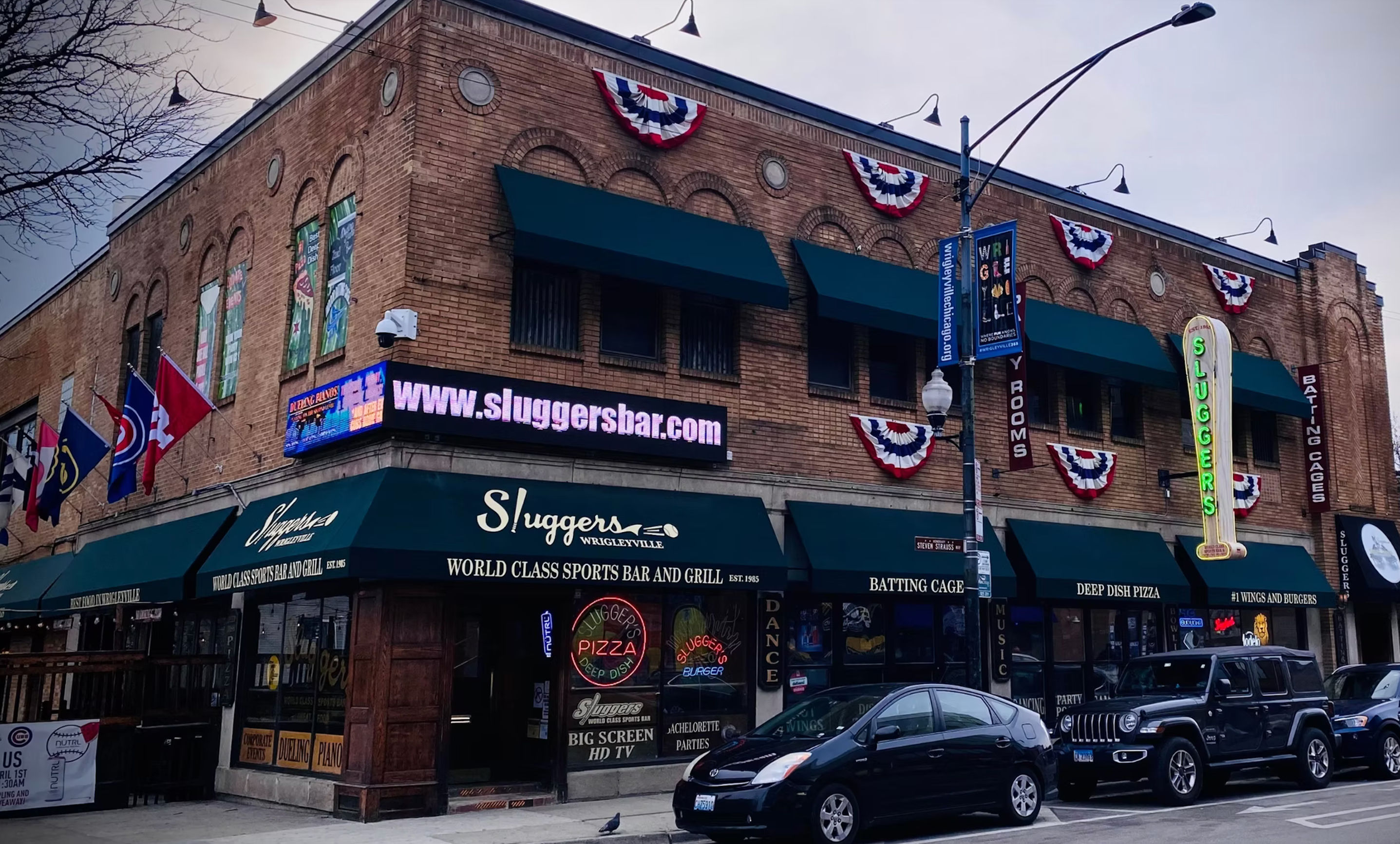 Brick corner sports bar and grill in Wrigleyville, Chicago with green awnings, patriotic bunting, neon vertical sign, flags and parked cars