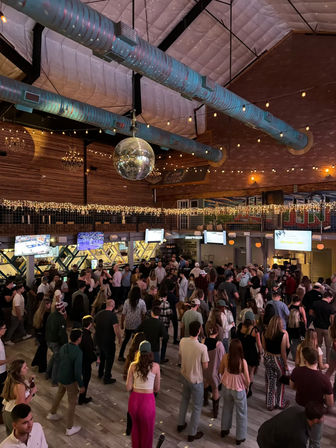 Urban nightlife venue with a mirrored disco ball, string lights and exposed ductwork over a brick-walled dance hall; large crowd dancing and mingling on a wooden floor with sports TVs above the bar.