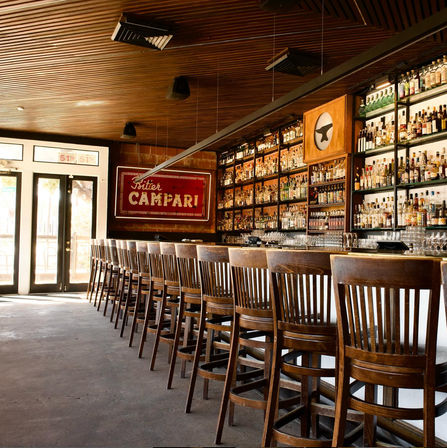 Sunlit cocktail bar interior with a long wooden counter and row of high wooden stools, backlit shelves full of liquor bottles and a vintage red wall sign.