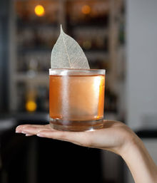 Hand holding a frosted rocks glass with an amber craft cocktail on the rocks, topped with a delicate skeleton leaf garnish, blurred bar background