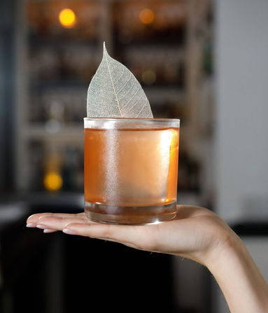 Hand holding a frosted rocks glass with an amber craft cocktail on the rocks, topped with a delicate skeleton leaf garnish, blurred bar background