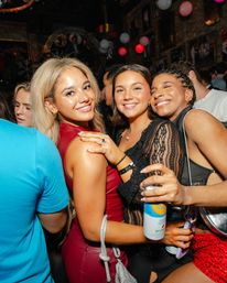 Three smiling women hugging at a crowded nightclub party with colorful balloons and lights overhead, one holding a canned drink — lively night out
