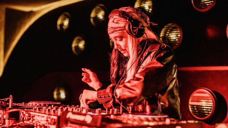 Female DJ wearing a bandana and headphones, mixing on a DJ controller in a red-lit nightclub with circular wall lights