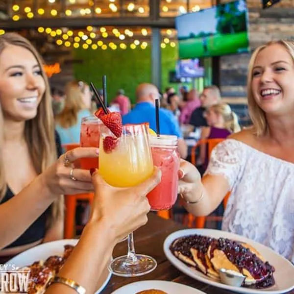Three friends toasting colorful brunch cocktails garnished with strawberries over pancakes and waffles at a lively downtown restaurant patio with string lights