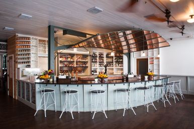 Bright coastal-style restaurant bar interior with a curved copper canopy over a pale blue paneled counter, row of white metal stools, dark wood floors and bottle-lined back shelves.