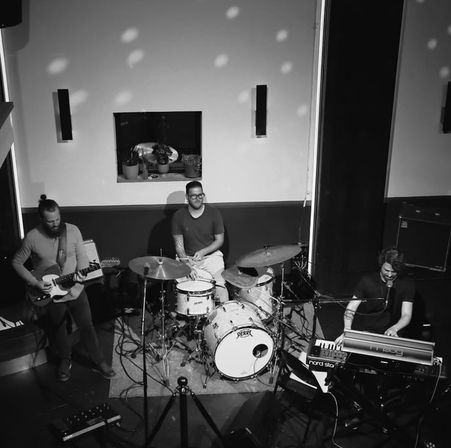 Black-and-white photo of a three-piece band performing live on a small indoor stage: guitarist, drummer and keyboardist/singer playing under scattered spotlights with a drum kit, keyboards and potted plants on a recessed windowsill behind them — intimate live music venue scene.