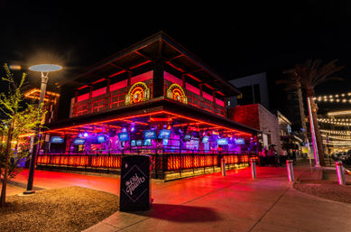Vibrant downtown nightlife scene with a neon-lit two-story bar and bustling outdoor patio glowing red and purple, string lights and palm trees lining a lively pedestrian street at night.