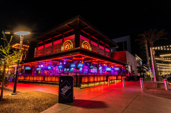 Vibrant downtown nightlife scene with a neon-lit two-story bar and bustling outdoor patio glowing red and purple, string lights and palm trees lining a lively pedestrian street at night.