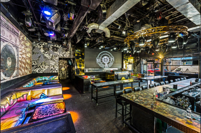 Vibrant urban nightclub interior with colorful LED-lit booths, long bar, stage with projection screen and DJ setup, exposed industrial ceiling and overhead lighting rig.