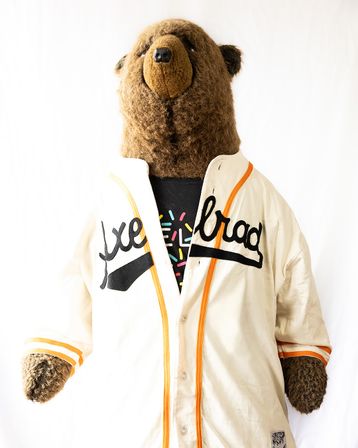 Plush bear mascot portrait wearing a cream vintage baseball-style jacket with orange trim and playful lettering against a white backdrop