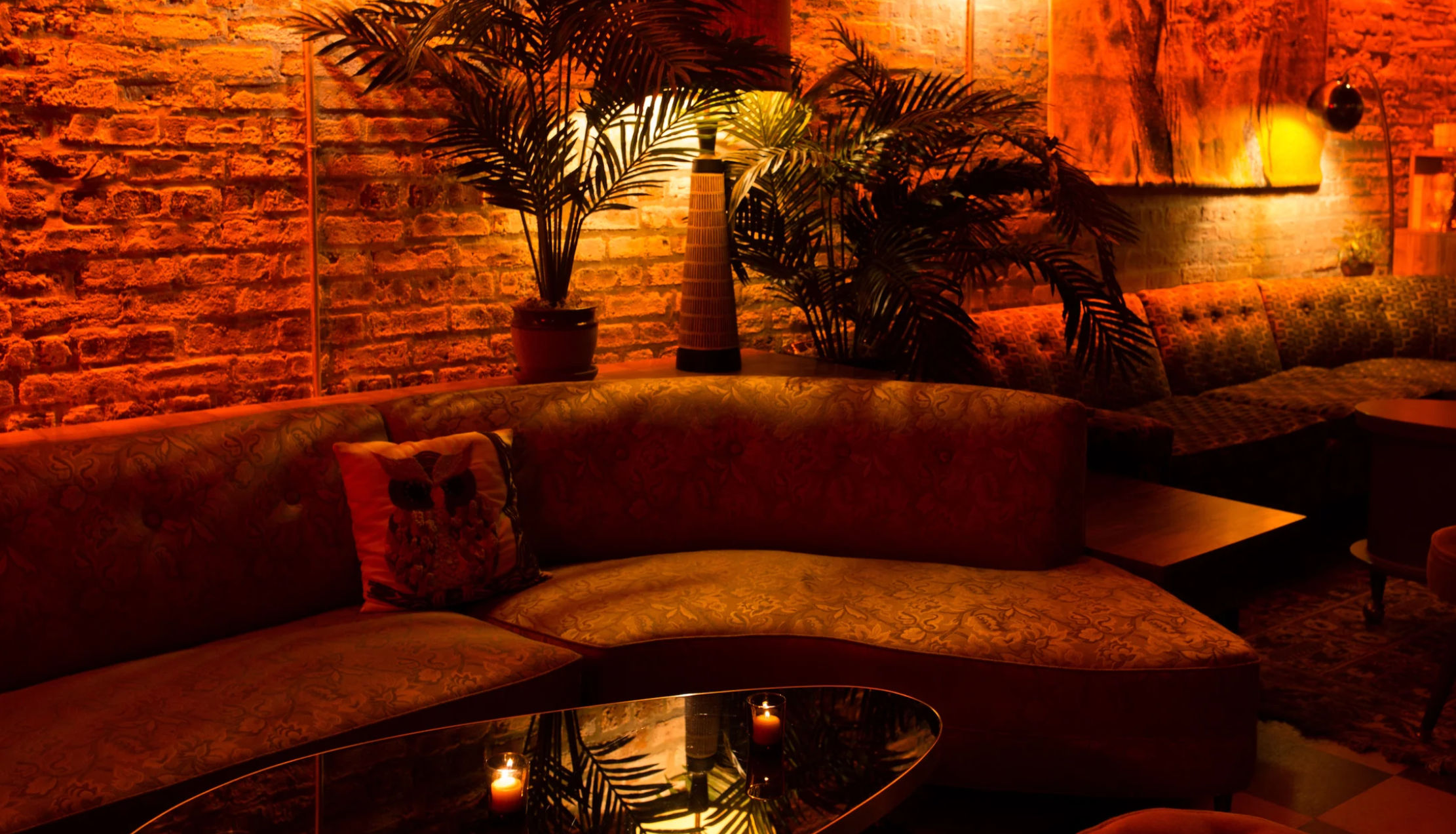 Cozy amber-lit lounge with a curved patterned sofa, exposed brick wall, potted palms, and a glass coffee table with flickering candles — intimate bar seating vibe.