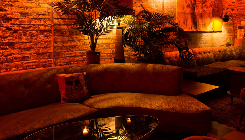 Cozy amber-lit lounge with a curved patterned sofa, exposed brick wall, potted palms, and a glass coffee table with flickering candles — intimate bar seating vibe.
