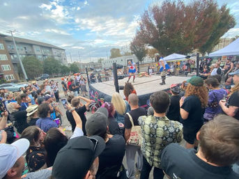 Outdoor street wrestling event in an urban neighborhood — lively crowd gathered around a raised ring watching costumed wrestlers compete, with tents, apartment buildings and autumn trees under a cloudy sky.