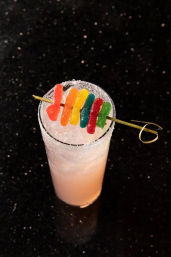 Pink tall cocktail in a sugar-rimmed glass with crushed ice, topped with a rainbow gummy worm skewer, pictured on a black speckled bar countertop