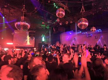 Packed nightclub dance floor under shimmering disco balls, neon red and purple lights, a lively crowd dancing, and a performer projected on a large screen.